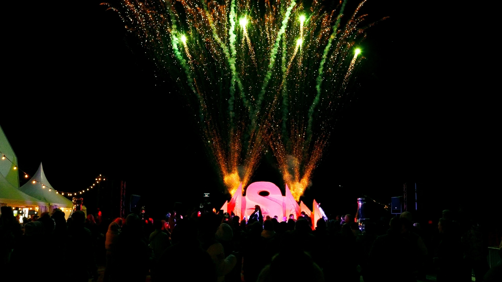 A festival crowd below a stage with green and orange fireworks at night.